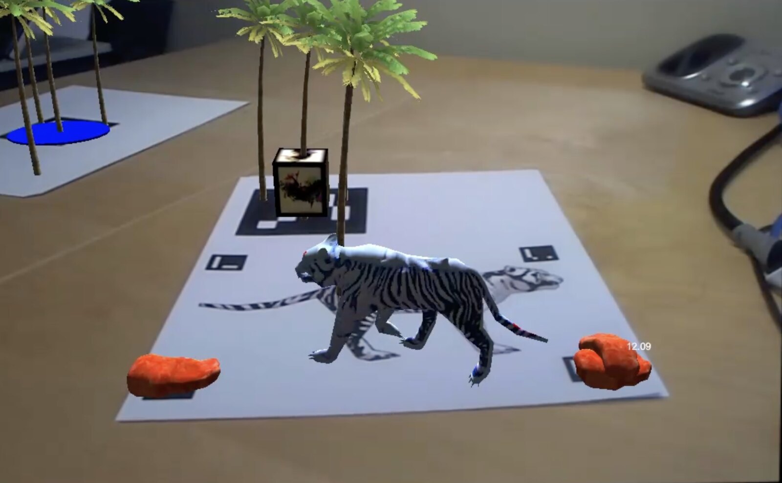 StickAR prototype: an uncoloured 3D tiger walking across a paper marker on a desk, palm trees behind it and a blue pond on the next sheet